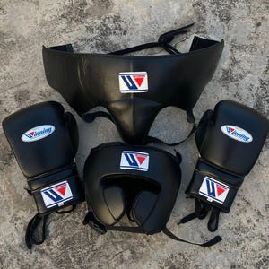 May include: A set of black boxing gear including two boxing gloves, a headgear, and a groin protector. Each piece features the Winning brand logo, which consists of a blue and red stylized letter W on a white rectangular patch.
