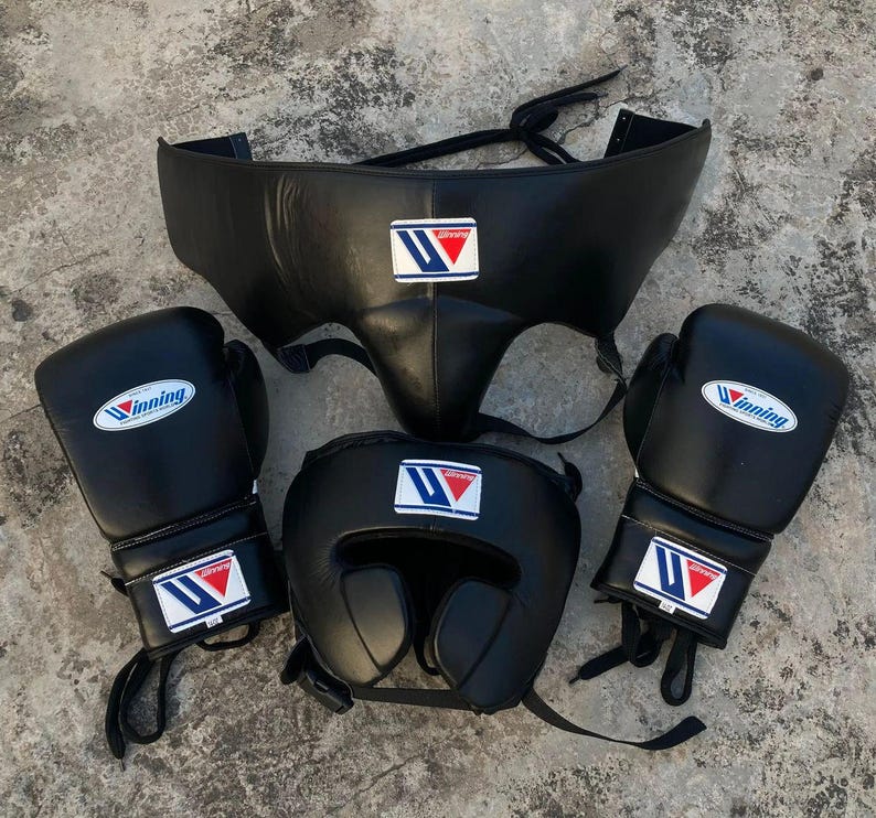 May include: A set of black boxing gear including two boxing gloves, a headgear, and a groin protector. Each piece features the Winning brand logo, which consists of a blue and red stylized letter W on a white rectangular patch.