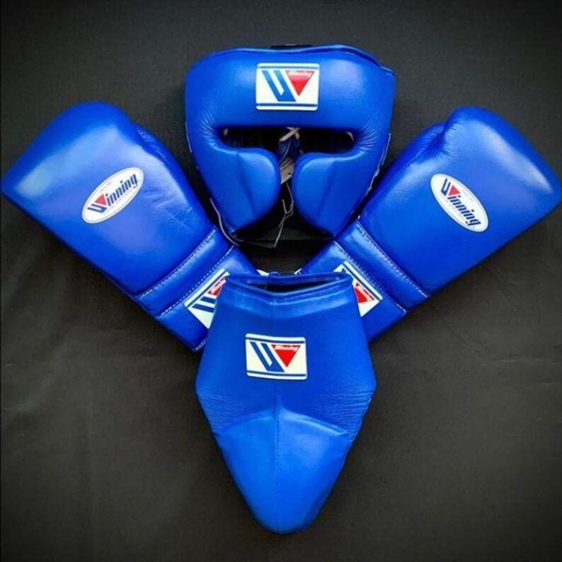 May include: A set of blue boxing equipment including two boxing gloves, a headgear, and a groin protector. Each piece features the white and red Winning logo. The items are constructed from smooth blue material with visible stitching details.