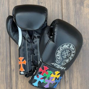 Puede incluir: Guantes de boxeo de cuero negro con costuras blancas y cierre con cordones. Los guantes presentan un logotipo blanco y diseños de cruces de colores. La palabra "ARTEAGA" está impresa en la muñeca.
