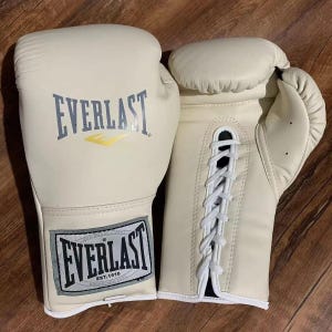 Puede incluir: Guantes de boxeo color crema con el logo "EVERLAST" en gris y amarillo. Los guantes están atados en la parte delantera con cordones blancos. Los guantes tienen apariencia de cuero, probablemente para entrenamiento o competición.