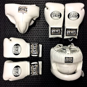 Puede incluir: Conjunto de equipo de boxeo blanco, que incluye guantes, casco y protector inguinal. Cada pieza presenta el logotipo "REYES" en negro y dorado. Los artículos están colocados sobre una superficie negra texturizada.