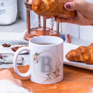 May include: White ceramic mug with a gold geometric design and floral accents. The mug features the letter "B" in a gray color. The mug is filled with a light brown liquid and is sitting on a wooden surface.