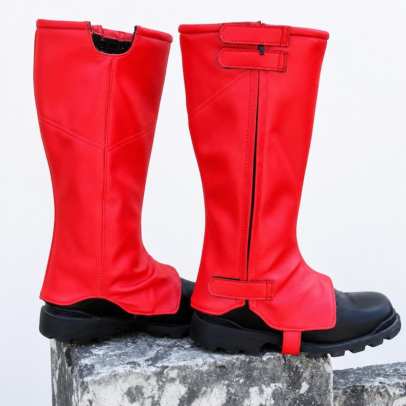 May include: A pair of bright red knee-high gaiters worn over black boots. The gaiters feature a vertical zipper closure along the side, secured with two adjustable hook-and-loop straps at the top and one strap at the base for a secure fit.