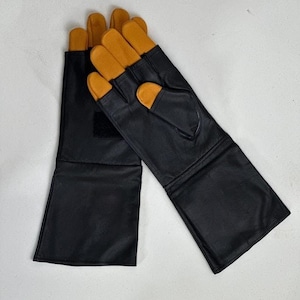 May include: A pair of long black leather gloves featuring tan leather fingertips and a thumb section. The gloves have an extended cuff design and a small black hook-and-loop fastener patch on the back of the wrist area for a secure fit.