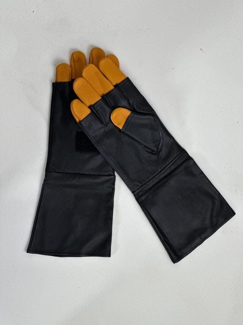 May include: A pair of long black leather gloves featuring tan leather fingertips and a thumb section. The gloves have an extended cuff design and a small black hook-and-loop fastener patch on the back of the wrist area for a secure fit.