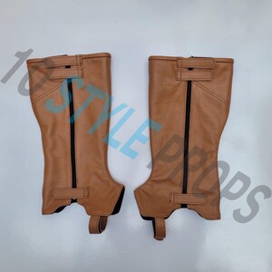May include: A pair of tan leather half chaps featuring vertical black zippers running down the center, adjustable hook-and-loop straps at the top and bottom, and a reinforced stirrup strap at the base for secure placement over footwear.