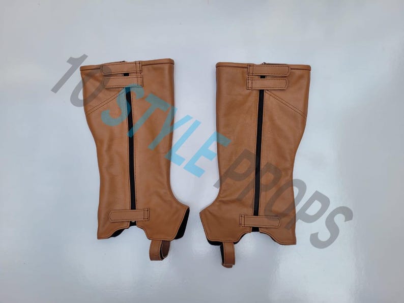 May include: A pair of tan leather half chaps featuring vertical black zippers running down the center, adjustable hook-and-loop straps at the top and bottom, and a reinforced stirrup strap at the base for secure placement over footwear.