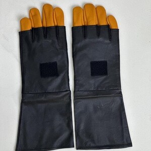 May include: A pair of fingerless gloves featuring long black leather cuffs and yellow leather fingers. Each glove has a square black hook-and-loop fastener patch centered on the back of the hand, extending from the wrist to the mid-forearm.