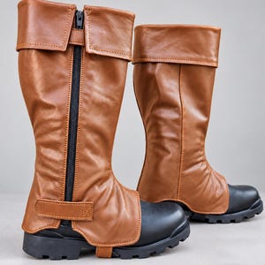 May include: A pair of brown leather-look boot covers featuring a folded cuff at the top, a vertical black zipper closure along the side, and a horizontal strap across the lower section. The covers are worn over black boots with thick lug soles.
