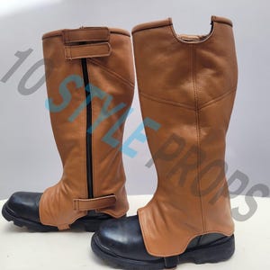May include: A pair of knee-high gaiters featuring a tan leather upper section with black vertical zippers and adjustable hook-and-loop straps, paired with black rounded-toe boots that have thick, lugged rubber soles and the text 10 STYLE PROPS.