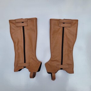 May include: A pair of tan equestrian half chaps featuring a vertical black zipper closure, two horizontal hook-and-loop fastener straps at the top, one horizontal strap at the bottom, and a stirrup strap extending from the base of each unit.
