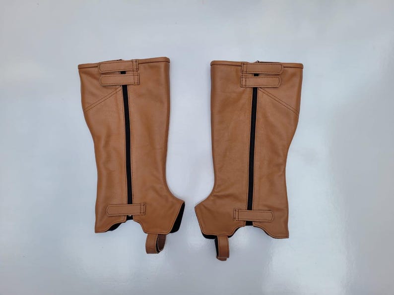 May include: A pair of tan equestrian half chaps featuring a vertical black zipper closure, two horizontal hook-and-loop fastener straps at the top, one horizontal strap at the bottom, and a stirrup strap extending from the base of each unit.