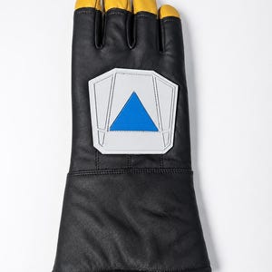 May include: A black leather gauntlet glove featuring yellow leather fingertips and a prominent white pentagonal patch on the back of the hand. The patch contains a centered blue triangle design with stitched detailing around the edges.