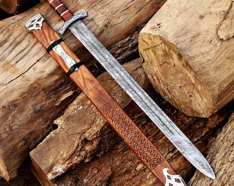 May include: A decorative sword with a patterned blade and a wooden handle and sheath. The handle is brown with silver accents. The sheath is also brown with a carved design. The sword is lying on a pile of wooden logs.