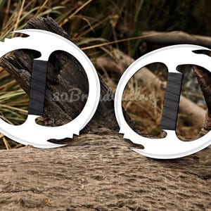 May include: Two white, circular throwing knives with black handles are displayed on a weathered log. The knives have a unique design with pointed edges and a central handle. The background features natural wood and foliage.