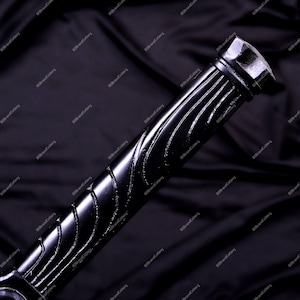 May include: Close-up of a black sword handle with a textured, ridged design. The handle has a glossy finish and a geometric end cap. The background is a dark, draped fabric.