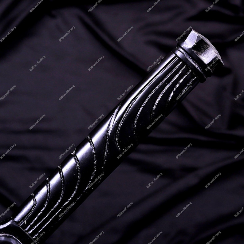 May include: Close-up of a black sword handle with a textured, ridged design. The handle has a glossy finish and a geometric end cap. The background is a dark, draped fabric.
