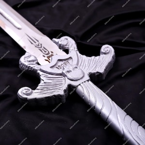 May include: A silver-colored sword with a detailed hilt and blade. The hilt features an ornate design with a skull-like emblem. The blade has a decorative etching. The sword is displayed against a black background.