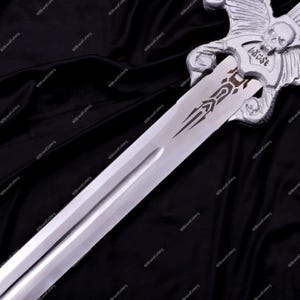 May include: A silver-colored sword with a skull and wing design on the hilt. The blade has a decorative etching. The word "b&uuml;st" is visible on the hilt. The sword is displayed against a dark background.