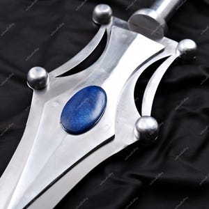 Puede incluir: Primer plano de una espada plateada con una hoja pulida y una piedra preciosa ovalada azul en el centro. La espada presenta detalles decorativos plateados y tachuelas plateadas redondeadas. El mango tambi&eacute;n es plateado.