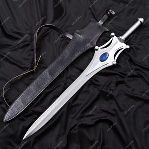 Puede incluir: Una espada plateada con una gema azul en la empu&ntilde;adura, junto a una vaina de cuero negro. La espada tiene un mango decorativo y una hoja larga y afilada. La vaina tiene una correa. La longitud de la espada es de aproximadamente 76 cm.