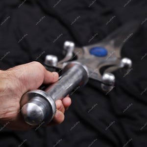 Puede incluir: Una espada plateada con una gema azul en el centro de la hoja. La empu&ntilde;adura y la guarda son detalladas. La espada se sostiene en una mano sobre un fondo negro.