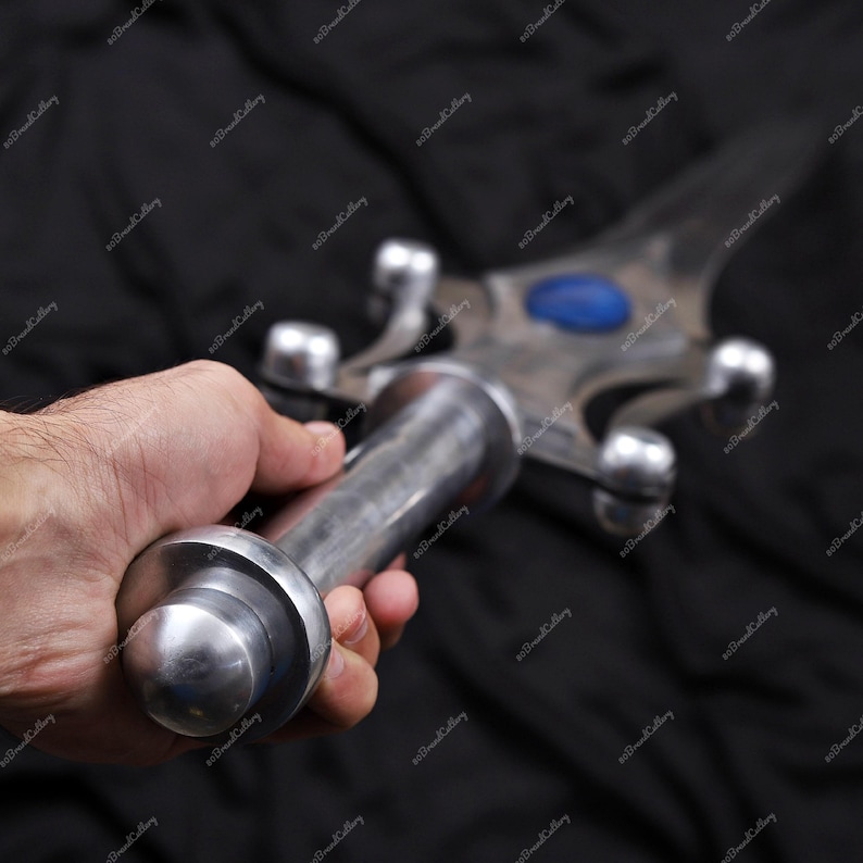Puede incluir: Una espada plateada con una gema azul en el centro de la hoja. La empu&ntilde;adura y la guarda son detalladas. La espada se sostiene en una mano sobre un fondo negro.