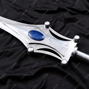Puede incluir: Primer plano de una espada plateada con una piedra ovalada azul en el centro de la hoja. La espada tiene un dise&ntilde;o decorativo con detalles plateados redondeados. El fondo es una tela oscura y texturizada.
