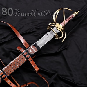 May include: A decorative sword with a patterned blade, gold-colored hilt, and a red and gold handle. The sword is paired with a brown leather sheath and straps. The text "80 Brand Cutlery" is in the upper left corner.