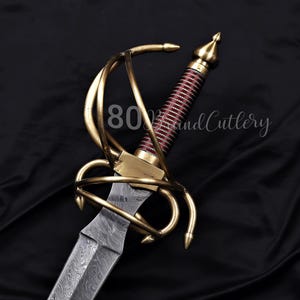 May include: A white ceramic mug with a black handle and the text "But First, Coffee" in a bold, black font. The mug is set against a blurred background, suggesting a cozy setting.