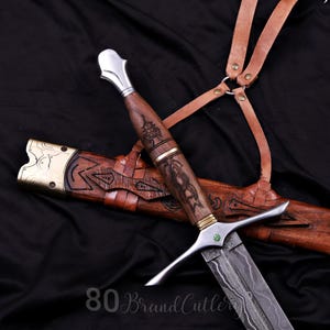 May include: A dagger with a Damascus steel blade, a wooden handle featuring intricate carvings, and a silver-toned pommel. The crossguard includes a small green stone. It is accompanied by a wooden scabbard with brass accents and leather straps.