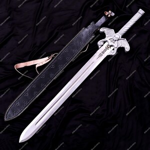 May include: A silver sword with a decorative hilt featuring a skull and wings design, resting on a black surface. The sword has a long, double-edged blade and is accompanied by a black leather sheath with a brown strap.