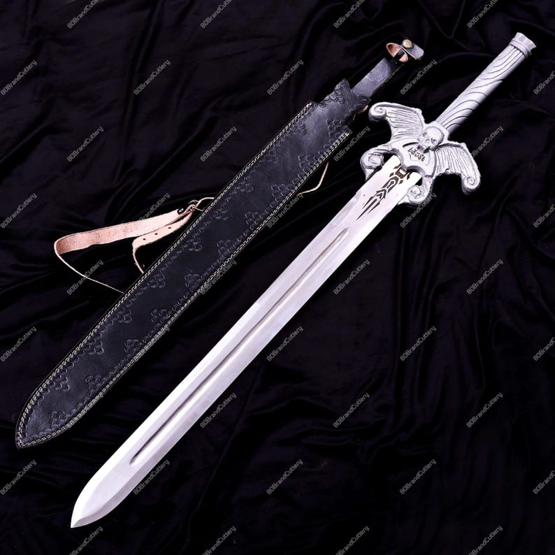 May include: A silver sword with a decorative hilt featuring a skull and wings design, resting on a black surface. The sword has a long, double-edged blade and is accompanied by a black leather sheath with a brown strap.