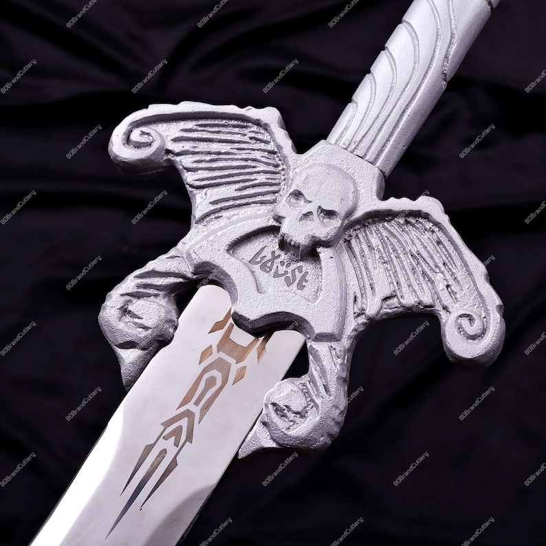 May include: A silver-colored sword with a skull and wing design on the hilt. The blade has a decorative pattern. The word "ASARA" is etched on the hilt. The sword is displayed against a black background.