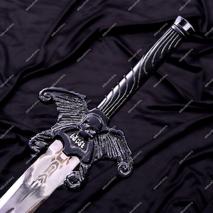 May include: A black fantasy sword with a silver blade. The hilt features a skull and wing design, with the word "BAST" etched on the guard. The blade has an etched design. The sword is displayed against a dark, textured background.