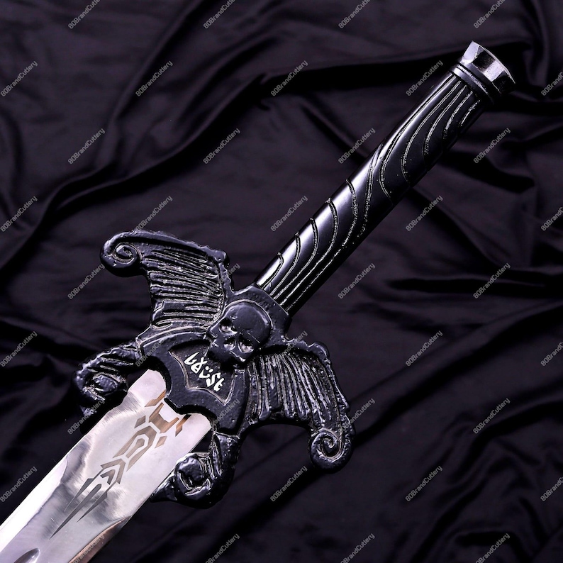 May include: A black fantasy sword with a silver blade. The hilt features a skull and wing design, with the word "BAST" etched on the guard. The blade has an etched design. The sword is displayed against a dark, textured background.