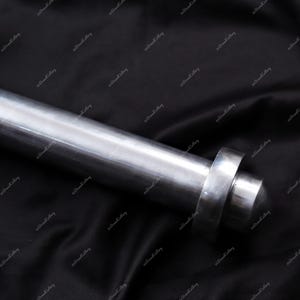 Puede incluir: Primer plano de un objeto met&aacute;lico cil&iacute;ndrico pulido, con un extremo redondeado y un detalle en forma de anillo. El objeto es plateado y parece estar hecho de acero o un metal similar. El fondo es una tela oscura y texturizada.