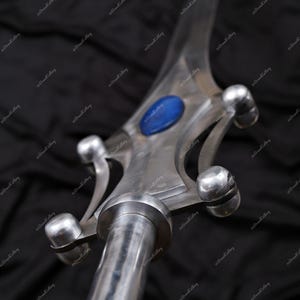 Puede incluir: Primer plano de una punta de lanza de metal plateado con una gema azul en el centro. La punta de lanza tiene un acabado pulido y detalles redondeados. La lanza es probablemente un art&iacute;culo decorativo o de colecci&oacute;n.
