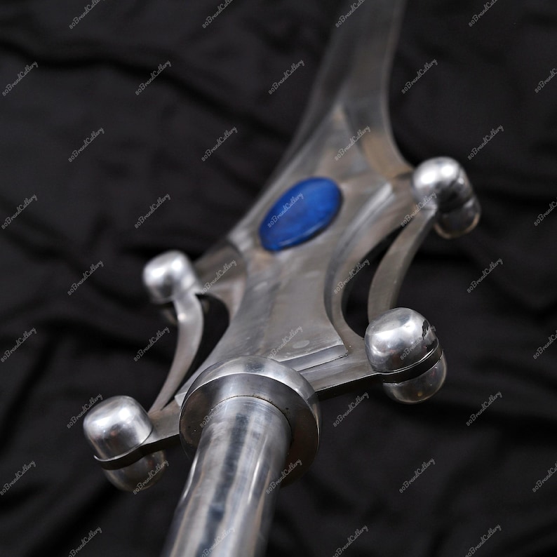 Puede incluir: Primer plano de una punta de lanza de metal plateado con una gema azul en el centro. La punta de lanza tiene un acabado pulido y detalles redondeados. La lanza es probablemente un art&iacute;culo decorativo o de colecci&oacute;n.