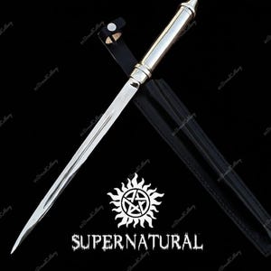 Puede incluir: Una daga plateada y negra con una funda de cuero. La daga tiene una hoja y un mango plateados, con una funda de cuero negro. La palabra "SUPERNATURAL" está impresa en blanco debajo de un símbolo de pentagrama blanco.
