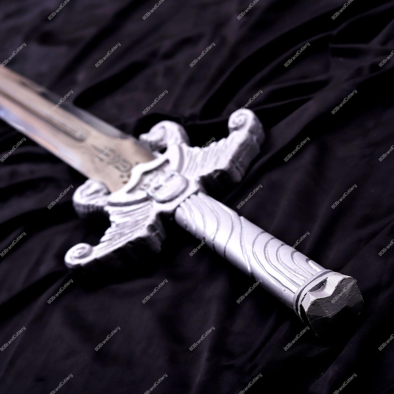 May include: A silver-colored sword with an ornate hilt and a detailed blade. The hilt features a decorative design with a shield-like emblem. The sword is set against a dark, textured background, highlighting its metallic sheen. A fantasy-themed item.