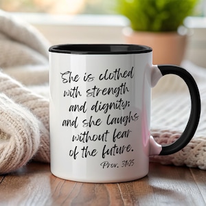Puede incluir: Taza de cerámica blanca con interior y asa negros. La taza presenta el texto "She is clothed with strength and dignity; and she laughs without fear of the future. Prov. 31:25" en escritura negra.