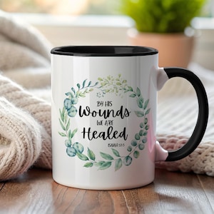 Puede incluir: Taza de cerámica blanca con interior y asa negros. La taza presenta el texto "By His Wounds We Are Healed" dentro de una corona de hojas verdes y bayas verde-azuladas. El texto "Isaiah 53:5" está debajo.