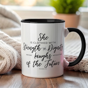 May include: White ceramic mug with a black interior and handle. The mug features the text "She is clothed with Strength and Dignity and she laughs of the Future" in black script. The Bible verse Proverbs 31:25 is also printed.