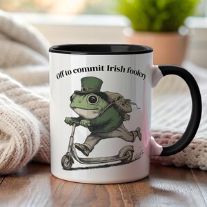 May include: White ceramic mug with a black interior and handle. The mug features a whimsical illustration of a frog riding a scooter, wearing a green hat and coat, with the text "Off to commit Irish foolery."
