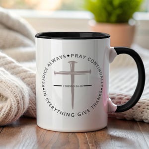 Puede incluir: Taza de cerámica blanca con interior y asa negros. La taza presenta un diseño de cruz con el texto "Dad gracias en todo, estad siempre alegres, orad sin cesar" y la referencia bíblica I Tes 5:16-18.