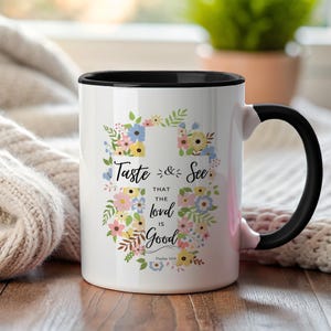 Puede incluir: Taza de cerámica blanca con interior y asa negros. La taza presenta un diseño de corona floral con el texto "Taste & See that the Lord is Good" en una fuente cursiva. El Salmo 34:8 está impreso en la parte inferior.