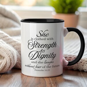 Puede incluir: Taza de cerámica blanca con interior y asa negros. La taza presenta el texto "She is clothed with Strength & Dignity and she laughs without fear of the future. Proverbs 31:25" en escritura negra.