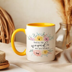 Puede incluir: Taza de cerámica blanca con asa e interior amarillos. La taza presenta ilustraciones florales en rosa, amarillo y verde, con el texto "And by His wounds we are healed." Un artículo decorativo para el hogar.